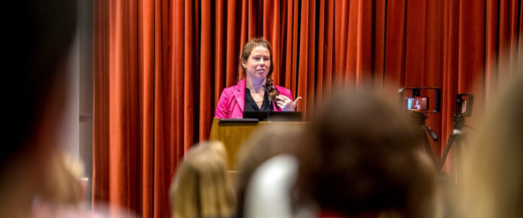 A woman behind a podium on stage giving a talk to to a room of people