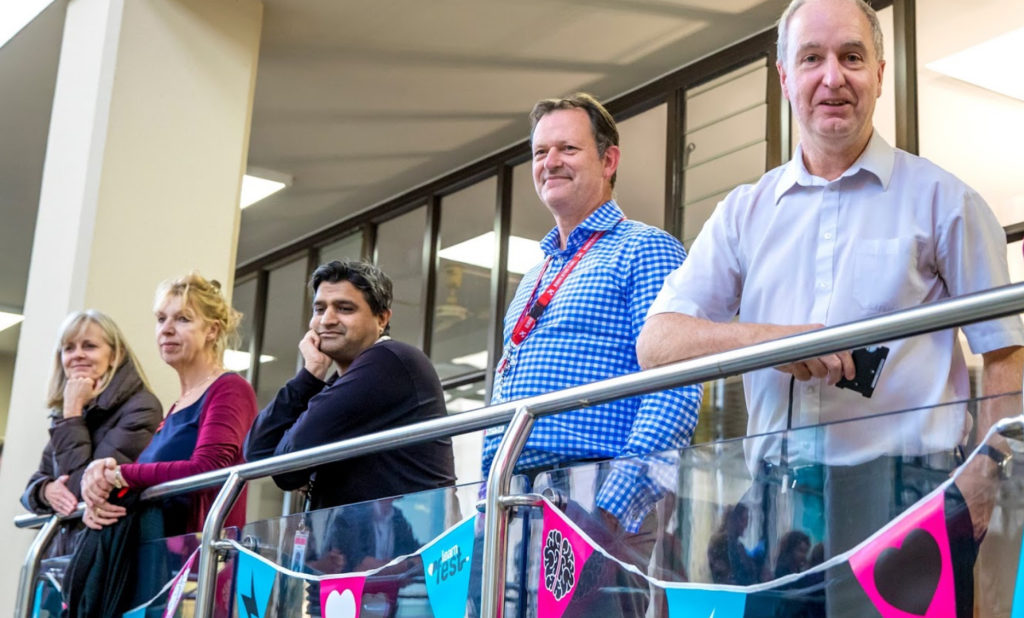 Two women ad three men leaning against a balcony rail watching a presentation